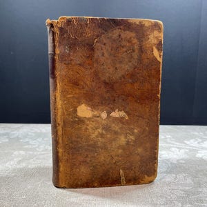 May include: A vintage, small, leather-bound book with a worn, distressed appearance. The cover is a rich brown with visible wear and tear, suggesting age and use. The spine shows signs of age, with a slightly tattered top edge. The book is resting on a patterned surface.