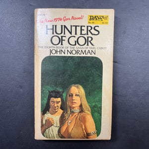 May include: A mass market paperback book titled Hunters of Gor by John Norman, the eighth book of the saga of Tarl Cabot. The cover features a green illustration of two figures with chains around their necks, with the text The New 1974 Gor Novel.