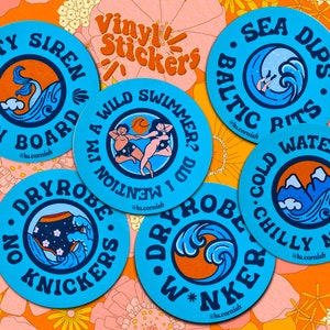 May include: Six round blue vinyl stickers with white text and illustrations. The stickers feature phrases like "Salty Siren on Board", "I'm a Wild Swimmer? Did I Mention", "Sea Dips. Baltic Rits", "On Dryrobe. No Knickers", "Water Dips. Cold Chilly Nips", and "Dryrobe W*Nker".