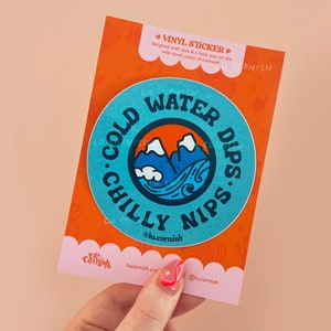 May include: A vinyl sticker with a blue circular design, featuring the text "COLD WATER DIPS. CHILLY NIPS." and a mountain and wave illustration. The sticker is on an orange card with text and a pink scalloped edge.
