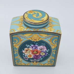 Könnte beinhalten: Eine quadratische Teedose aus Keramik mit floralem Design. Die Dose ist mit goldenen Verzierungen und einem floralen Muster auf hellblauem Hintergrund verziert. Der Deckel ist rund und hat eine goldene Verzierung mit einem floralen Muster.