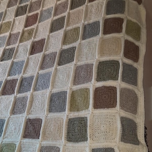 Könnte beinhalten: Eine handgefertigte Häkeldecke mit einem Patchwork-Design. Die Decke besteht aus zahlreichen quadratischen Blöcken in verschiedenen neutralen Farben, darunter Creme, Grau, Braun und Grün. Die Textur der Decke ist sichtbar.