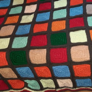 Könnte beinhalten: Eine handgefertigte Häkeldecke mit einem Gitter aus bunten quadratischen Flicken in den Farben Rot, Orange, Blau, Grün, Beige und Grau, die durch einen dunkelbraunen Garnrand verbunden sind, der einen deutlichen Rahmen um jedes einzelne Quadrat bildet.
