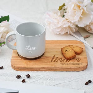 May include: A light grey ceramic mug with the name "Lisa" engraved on it, sitting on a wooden tray with the text "Lisa's Tea & Biscuits". Two light brown biscuits are on the tray.