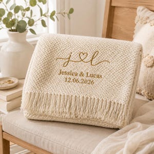Könnte beinhalten: Eine cremefarbene Strickdecke mit strukturiertem Muster und Fransen. Die Decke ist mit dem goldfarbenen Text "j & l" über einem Herzsymbol bestickt, gefolgt von den Namen "Jessica & Lucas" und dem Datum "12.06.2026".