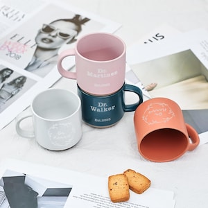 May include: Four ceramic mugs in shades of pink, teal, and peach, with white text and handles. The mugs are stacked and arranged on a surface with magazine pages and cookies. The text on the mugs reads "Dr. Martinez", "Dr. Walker", and "Dr. Harris".