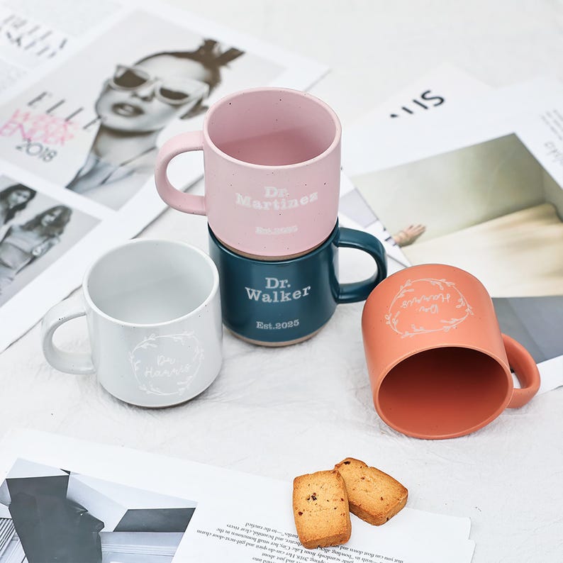 May include: Four ceramic mugs in shades of pink, teal, and peach, with white text and handles. The mugs are stacked and arranged on a surface with magazine pages and cookies. The text on the mugs reads "Dr. Martinez", "Dr. Walker", and "Dr. Harris".