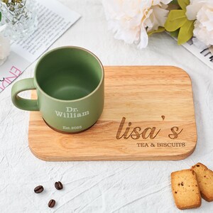 May include: A green ceramic mug with the text "Dr. William Est.2025" sits on a wooden serving board. The board is engraved with "lisa's TEA & BISCUITS". Two biscuits and coffee beans are also present.
