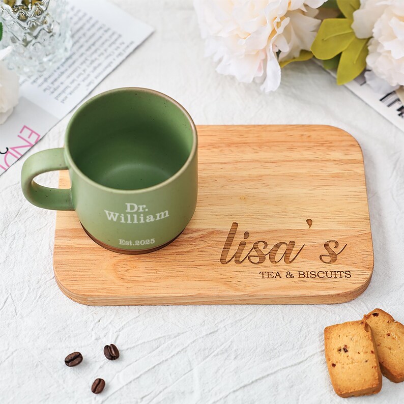 May include: A green ceramic mug with the text "Dr. William Est.2025" sits on a wooden serving board. The board is engraved with "lisa's TEA & BISCUITS". Two biscuits and coffee beans are also present.