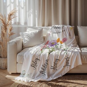 May include: A white blanket with a floral design, draped over a cream sofa. The blanket features pink, purple, and yellow flowers, with the words "Grandma's Garden, Love Grows Here" and the names Riley, Addison, Henry, and James.