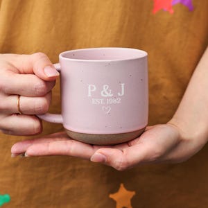 Puede incluir: Una taza de cerámica rosa con un acabado moteado. La taza tiene un asa blanca y una inscripción blanca que dice "P&J EST. 1982" con un pequeño símbolo de corazón.