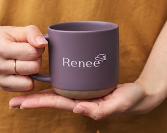 Op de afbeelding: Een paarse keramische mok met een gespikkelde afwerking en een bruine basis. De mok heeft de naam "Renee" in witte letters, samen met een kleine kerstmuts-afbeelding. De mok wordt in de handen van een persoon gehouden.