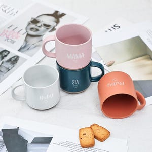 Pode incluir: Três canecas de cerâmica com nomes e cores diferentes. As canecas estão empilhadas uma em cima da outra. A caneca de cima é rosa com bolinhas brancas e o nome "MAMA" escrito nela. A caneca do meio é azul-esverdeada com o nome "SIA" escrito nela. A caneca de baixo é branca com o nome "Ruby" escrito nela. As canecas estão sobre uma superfície branca com dois biscoitos ao lado.