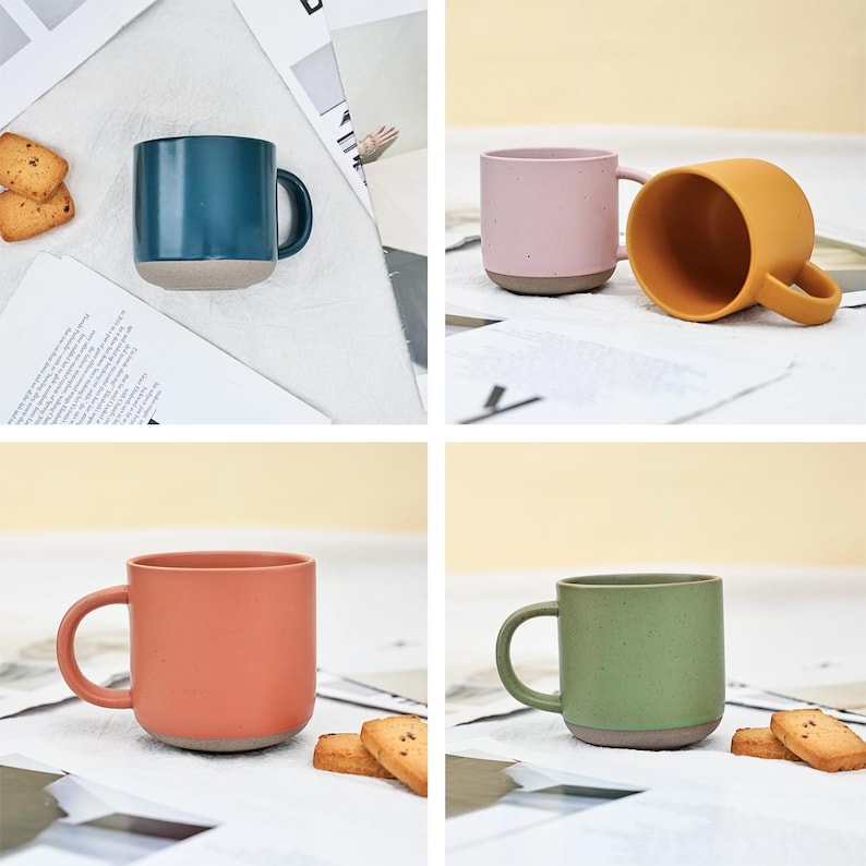 May include: A collection of ceramic mugs in various colors: teal, pink, orange, and green. Each mug has a contrasting gray base and a curved handle. The mugs are displayed on a white surface with scattered paper and cookies.