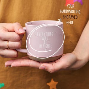 Può includere: Una tazza in ceramica rosa con una finitura macchiettata. La tazza ha un manico e dice "Everything will be alright." in lettere bianche. La tazza è tenuta nella mano di una persona.