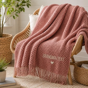 Op de afbeelding: Een oudroze gebreide plaid met een textuur van noppen en franjes. De deken is voorzien van de geborduurde tekst "GRANDMA/TEXT" in een lichte kleur, vergezeld van een klein geborduurd hartsymbool onder de belettering.