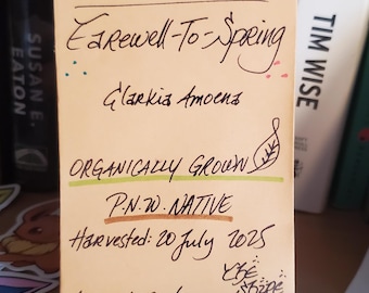 May include: A rectangular light orange paper label with handwritten black ink text reading Godetia Farewell-To-Spring Clarkia Amoena, Organically Grown, P.N.W. Native, Harvested: 20 July 2025, Approx. 100 seeds, Weight: 1 gram, and The Shire Life.