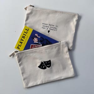 May include: Two off-white canvas zipper pouches. One pouch has the text "where better to escape trouble than a theatre?" and a heart. The other pouch has black theatre masks. A yellow Playbill is partially visible.