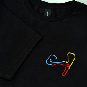 Könnte beinhalten: Ein schwarzes T-Shirt mit kurzen Ärmeln und einer gestickten Grafik einer Rennstrecke auf der Brust. Der Streckenverlauf ist in hellblauen, gelben und roten Garnsegmenten gestickt, ergänzt durch ein kleines schwarz-weiß kariertes Flaggendetail.