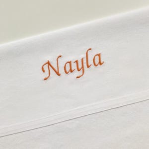 Può includere: Una superficie tessile bianca che presenta il nome Nayla ricamato in corsivo con filo arancione. Il tessuto ha una consistenza liscia e pulita con una cucitura orizzontale che attraversa la parte inferiore della composizione.