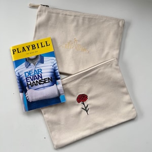 May include: Two beige canvas zipper pouches, one with a yellow embroidered skyline and the other with a red flower. A "Dear Evan Hansen" Playbill is next to the pouches.