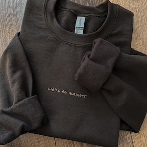 以下が含まれることがあります： 金色の糸で「WE'LL BE ALRIGHT」と刺繍された濃い茶色のスウェットシャツ。スウェットシャツは折りたたまれ、袖口は折り返されています。Gildan Heavy Blendのタグが見えます。