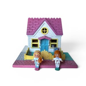Puede incluir: Una casa de muñecas en miniatura con techo rosa, paredes blancas y puerta azul. Dos pequeñas muñecas están de pie frente a la casa. La casa tiene ventanas amarillas y una pequeña ventana circular amarilla. La base es rosa y gris.