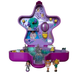 Può includere: Un playset giocattolo a forma di stella viola con un design aperto, con personaggi in miniatura e dettagli interni. Il playset include una mongolfiera, un orologio e altri elementi decorativi. Tre piccole bambole sono di fronte al playset.