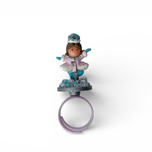May include: A ring with a small figurine of a girl in winter attire. The figurine wears a pink coat, blue gloves, and a blue hat. The ring itself is a light purple colour.