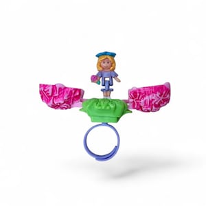 May include: A vintage Polly Pocket ring toy. It features a miniature doll with blonde hair and a blue hat. The toy has a green base, pink floral accents, and a blue ring. The doll is holding a small bouquet of flowers.