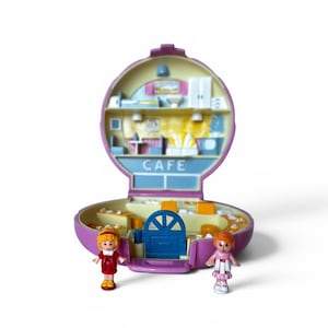 May include: A vintage, open, purple Polly Pocket cafe playset with two miniature figures. The cafe has a blue door, yellow tables, and the word "CAFE" in blue. The figures are wearing dresses and standing in front of the cafe.