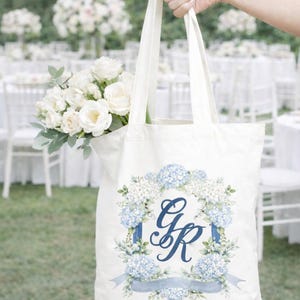Woman holding a tote bag with a personalized watercolor wedding crest monogram in blue florals at an outdoor wedding venue.