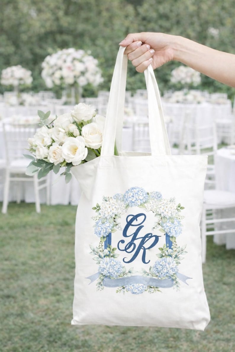 Woman holding a tote bag with a personalized watercolor wedding crest monogram in blue florals at an outdoor wedding venue.