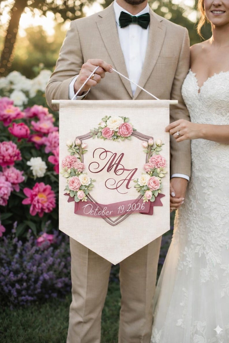 Bride and groom holding a personalized wedding banner with a watercolor crest monogram, pink florals and wedding date.