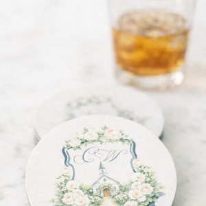 Round white coaster with custom watercolor wedding crest monogram featuring soft florals, initials and chapel illustration.