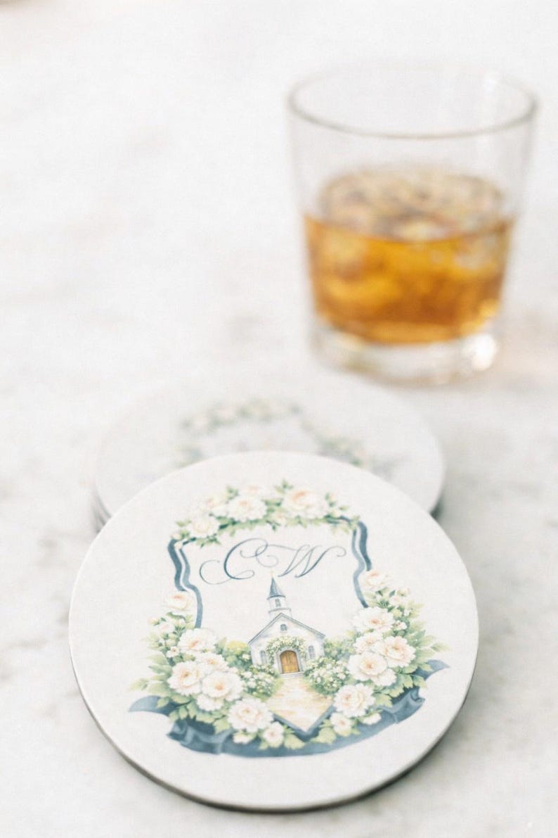 Round white coaster with custom watercolor wedding crest monogram featuring soft florals, initials and chapel illustration.