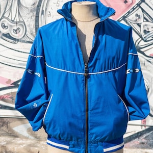 Op de afbeelding: Een blauwe windbreaker-jas met witte piping en een ritssluiting. De jas heeft een opstaande kraag en een geribbelde zoom en manchetten.
