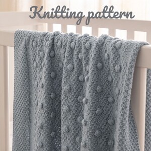 May include: A light blue knitted blanket with a textured bobble design draped over a white crib. The blanket has a soft, cozy appearance. The words "Knitting pattern" are written in a cursive font above the blanket.