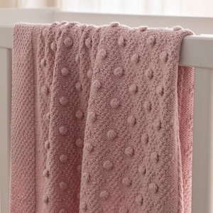 May include: A soft, pink knitted blanket with a textured bobble design, draped over a white crib. The blanket's pattern is visible, showcasing the detailed knitwork. The overall aesthetic is cozy and inviting, perfect for a nursery.