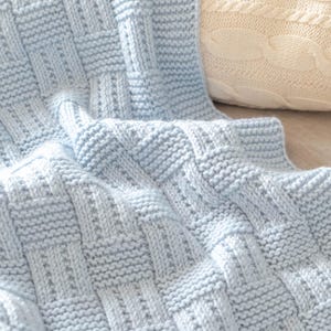 Puede incluir: Una manta de punto azul claro con un patrón de tejido de cesta. La manta está hecha de hilo suave y tiene una superficie texturizada. Un cojín crema de punto trenzado está en el fondo.