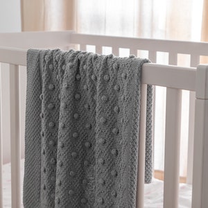 Pu&ograve; includere: Una coperta grigia lavorata a maglia con un motivo a pois strutturato, drappeggiata sul lato di una culla bianca. La consistenza morbida e il colore neutro della coperta suggeriscono un articolo accogliente e confortevole per la cameretta.