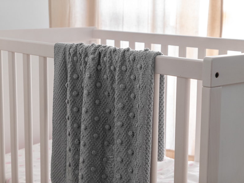 Pu&ograve; includere: Una coperta grigia lavorata a maglia con un motivo a pois strutturato, drappeggiata sul lato di una culla bianca. La consistenza morbida e il colore neutro della coperta suggeriscono un articolo accogliente e confortevole per la cameretta.