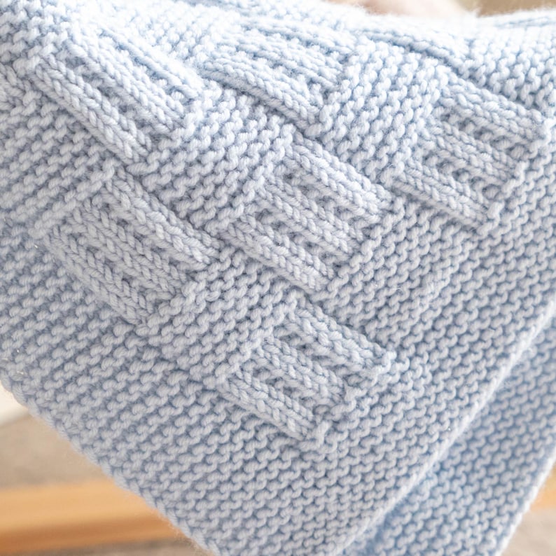 May include: A close-up of a light blue knitted blanket. The blanket features a textured, woven design in the center, with a smooth, knitted stitch pattern surrounding it. The blanket is folded over, showing the texture and depth of the knit.