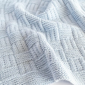 May include: A light blue knitted blanket with a basketweave pattern. The blanket is made of soft yarn and has a textured surface. The weave creates a geometric design, perfect for home decor.