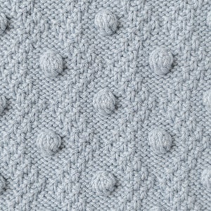 Pu&ograve; includere: Primo piano di un tessuto a maglia azzurro chiaro con un motivo strutturato. Il tessuto presenta un design ripetuto di pompon sferici in rilievo, uniformemente distanziati sulla superficie, creando una texture tattile e visivamente interessante. La maglia ha un aspetto sottile e morbido.