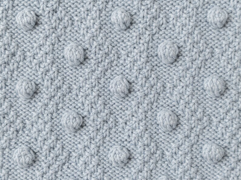 Pu&ograve; includere: Primo piano di un tessuto a maglia azzurro chiaro con un motivo strutturato. Il tessuto presenta un design ripetuto di pompon sferici in rilievo, uniformemente distanziati sulla superficie, creando una texture tattile e visivamente interessante. La maglia ha un aspetto sottile e morbido.