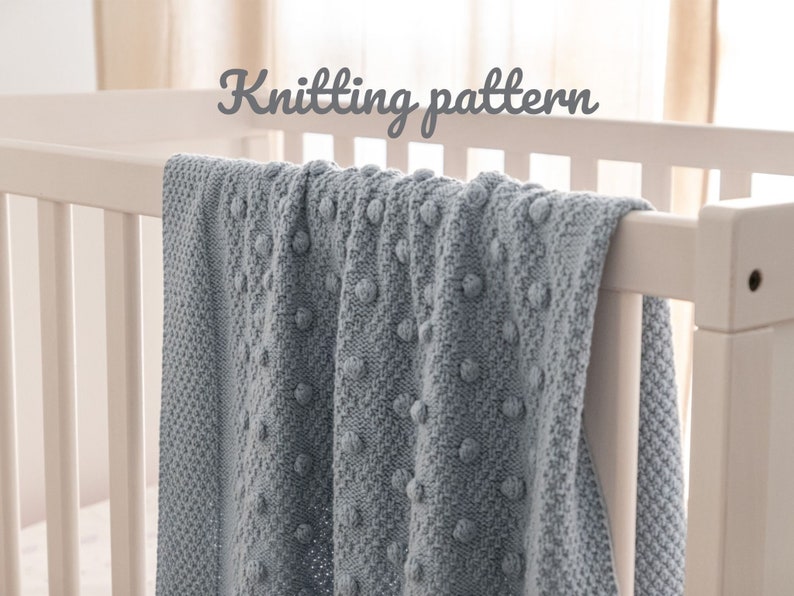 May include: A light blue knitted blanket with a textured bobble design draped over a white crib. The blanket has a soft, cozy appearance. The words "Knitting pattern" are written in a cursive font above the blanket.