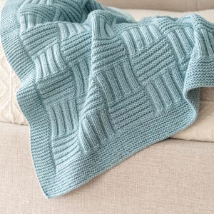 May include: A light blue knitted blanket featuring a textured basketweave pattern composed of alternating vertical and horizontal ribbed blocks, finished with a simple garter stitch border along the edges of the fabric.