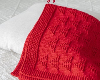 Può includere: Una coperta di maglia rossa con un motivo geometrico. La coperta è piegata e appoggiata su una superficie bianca. "Knitting pattern" è scritto in nero nella parte superiore dell'immagine.