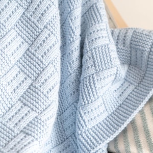 May include: A light blue knitted blanket with a textured, woven pattern. The blanket is folded, revealing the intricate stitch work and a border. The soft, cozy blanket is draped over a chair, suggesting comfort and warmth.